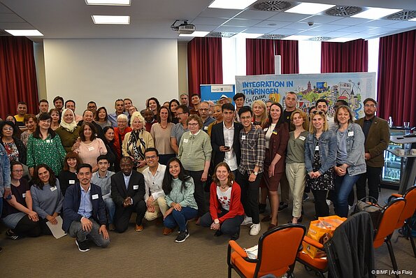 Foto MO VA: Gruppenfoto viele Menschen kniend und stehend vor einer Leinwand, einem Wimmelbild und einem Rollup in einem Raum im Vordergrund ein paar Stühle