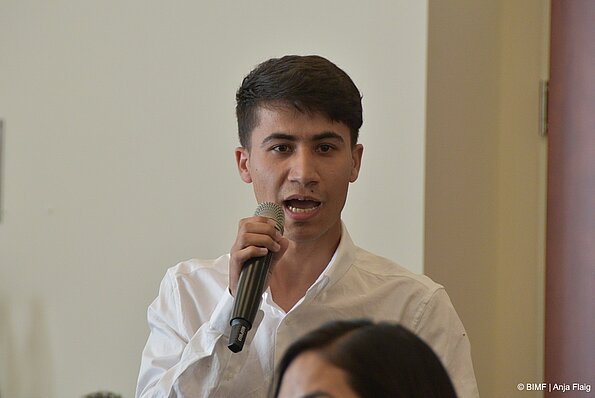 Foto MO VA: ein Mann sprechend in ein Mikrofon, ein Kopf unscharf unten im Bild