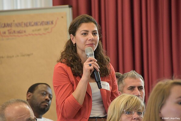 Foto MO VA:eine Frau stehend mit einem Mikrofon in der Hand im Hintergrund Menschen sitzend und ein Plakat