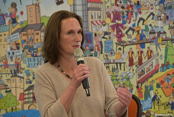 Foto MO VA: Mirjam Kruppa, Thüringer Beauftragte für Integration, Migration und Flüchtlinge stehend vor einem Wimmelbild mit Mikrofon