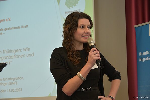 Foto MO VA: eine Frau stehend mit einem Mikrofon in der Hand im Hintergrund eine Prästentation mit Logo und Rollup
