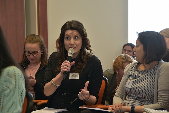 Foto MO VA: eine Frau sitzend spricht in ein Mikrofon in der Hand, drumherum weitere Personen sitzend