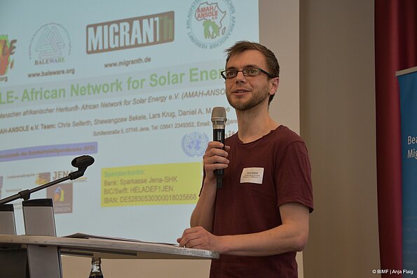 Foto MO VA: ein Mann mit Brille stehend vor einem Rednerpult mit einem Mikrofon in der Hand, im Hintergrund eine Präsentation mit Text und Logo