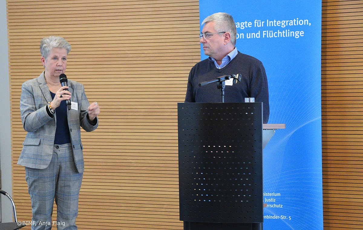 Ehrenamtsveranstaltung 20.11.2024, 2 Personen stehend, linke Person hält das Mikrofon in der Hand, rechte Person steht vor dem Rednerpult, im Hintergrund Holzlamellen und Rollup Thüringer Beauftragte für Integration, Migration und Flüchtlinge in blau mit weißer Schrift