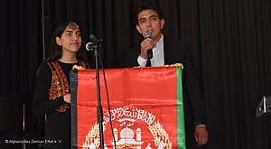 2 Personen (eine Frau und ein Mann) stehend am Mikrofon hinter einem Stehpult, eine Flagge