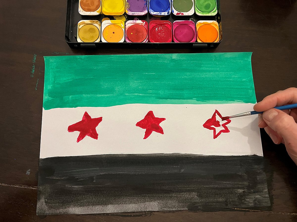 Wasserfarbkasten und Hand, die eine Syrien-Flagge malt: Trikolore in Grün-weiß-schwarz mit drei roten Sternen im mittleren weißen Balken.