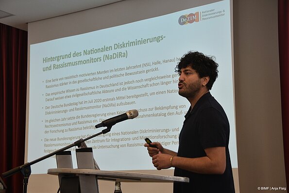 Foto MO VA: ein Mann stehend vor einem Pult mit Mikrofon, im Hintergrund eine Power-Point Präsentation mit Text