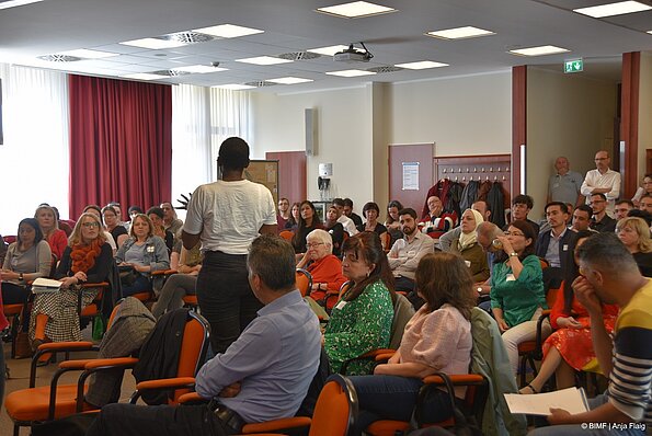 Foto MO VA: viele Menschen sitzend in einem Raum, eine Frau stehend mit dem Rücken zum Fotograf, im Hintergrund 2 stehende Personen
