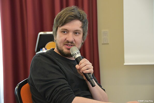 Foto MO VA: ein Mann sitzend mit Bart und Mikrofon in der Hand, sprechend