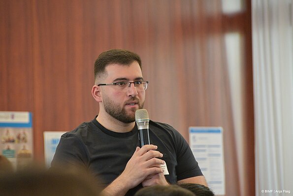 Foto MO VA: ein Mann mit einem Mikrofon in der Hand stehend vor einer Wand mit Plakaten