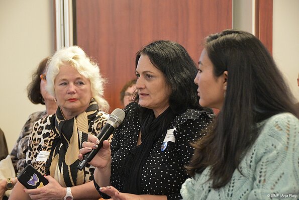 Foto MO VA: drei Frauen stehend, die Frau in der Mitte hält das Mikrofon, im Hintergrund Menschen