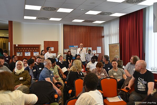 Foto MO VA: viele Menschen sitzend und schauend zu einem Mann stehend mit Mikrofon in der Hand in einem Raum