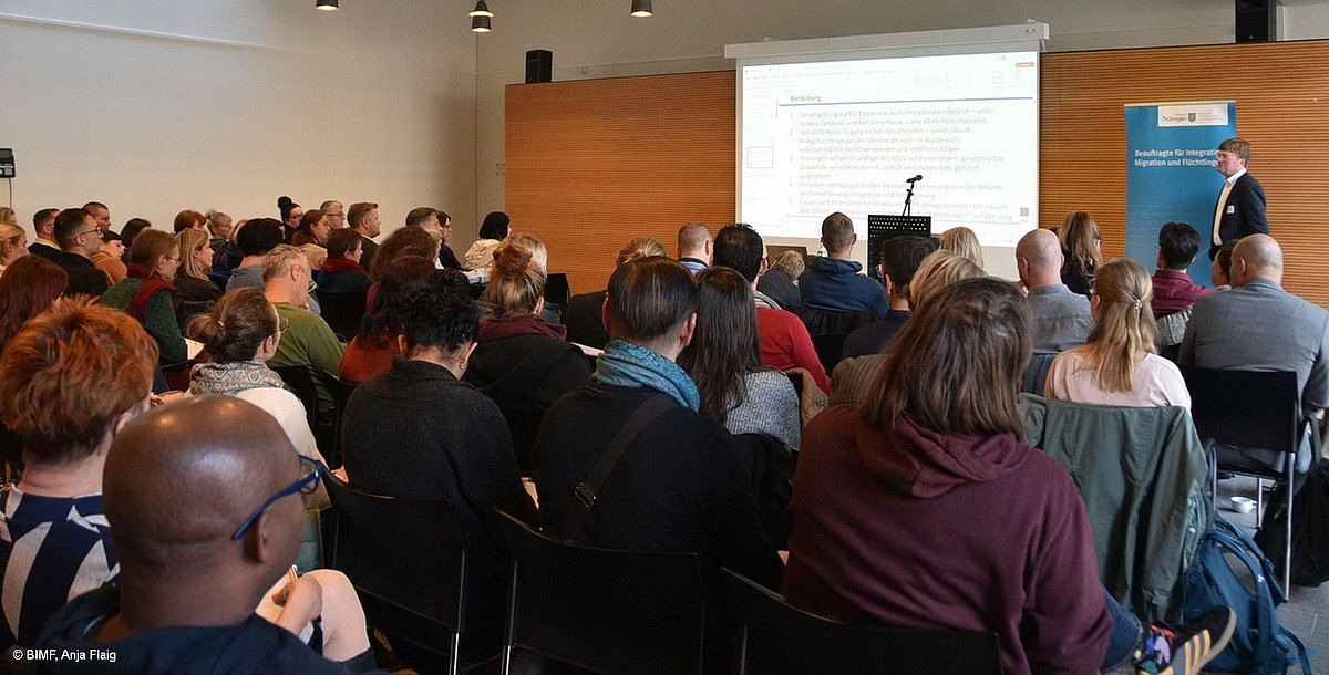 Publikum von hinten, viele verschiedene Menschen sitzend und schauend zum Referenten und Prästentation