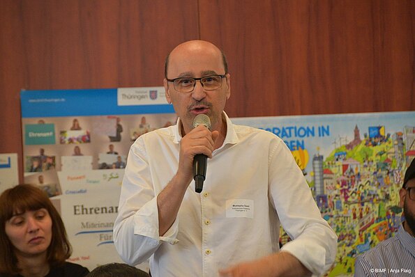 Foto MO VA: ein Mann mit Brille und Mikrofon in der Hand stehend vor 2 Plakaten, Menschen sitzend