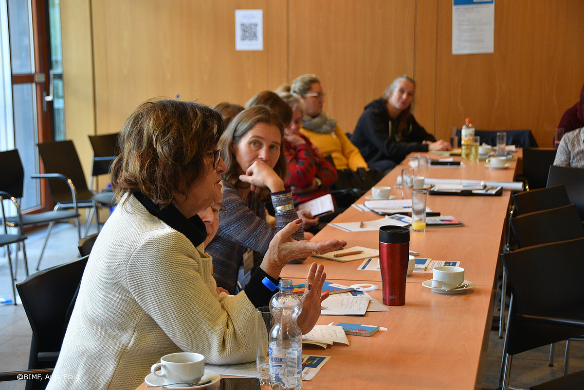 Ehrenamtsveranstaltung 20.11.2024 Publikum seitlich sitzend am Tisch, mehrere Personen eine Frau schaut direkt in die Kamera. Auf dem Tisch liegen Papiere für Mitschriften, Gläser und Tassen