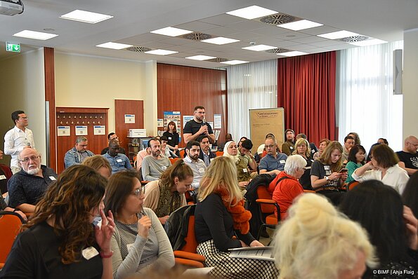 Foto MO VA: viele Menschen hauptsächlich sitzend, 3 stehend, ein Mann mit Mikrofon in der Hand