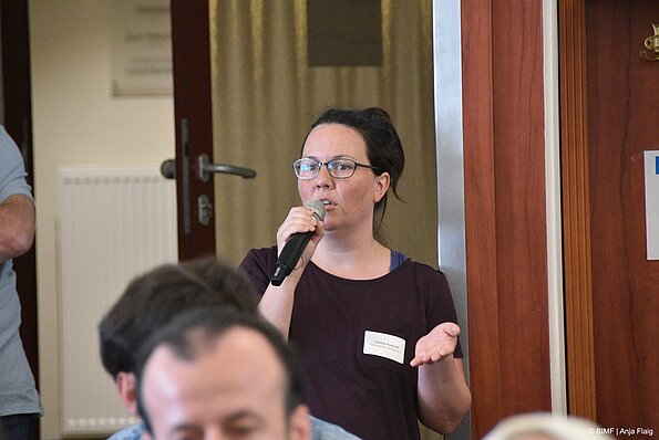 Foto MO VA: eine Frau stehend mit Brille und einem Mikrofon in der Hand, verschiedene Menschen unscharf im unteren Bildrand