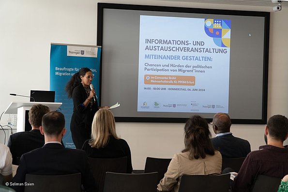 Veranstaltung politische Teilhabe 06.06.2024 - Frau stehend vor Leinwand, mit einem Mikrofon, im Vordergrund Publikum sitzend