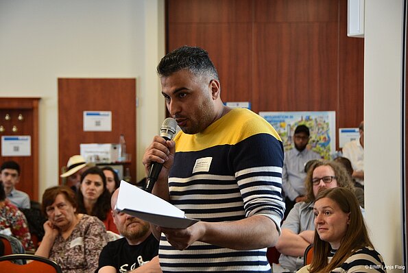 Foto MO VA: ein Mann stehend mit Mikrofon in der Hand, im Hintergrund Menschen sitzend