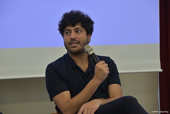 Foto MO VA: ein Mann sitzend mit einem Mikrofon in der Hand, im Hintergrund eine Leinwand mit blauem Bild