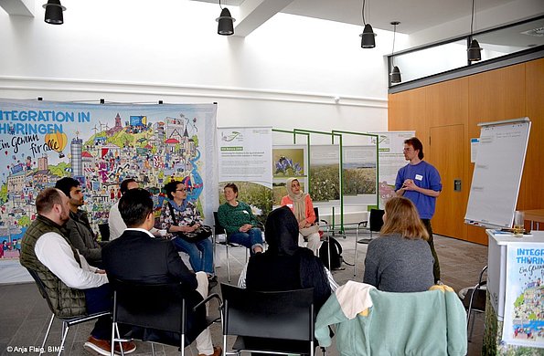 MO VA 17.05.2025 Gesprächsrunde, mehrere Personen sitzend im Halbkreis, eine Person stehend, im Hintergrund ein Wimmelbild