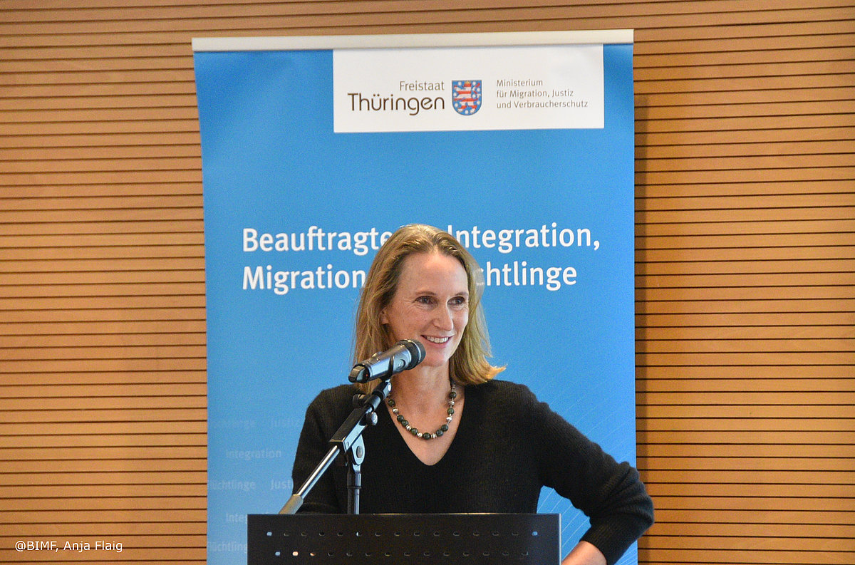 Veranstaltung "Engagiert für Vielfalt: Ehrenamt in der Integrationsarbeit in Thüringen - In Gegenwart und Zukunft" Mirjam Kruppa, Beauftragte, stehend vor dem Mikrofon, im Hintergrund Rollup der Beauftragten