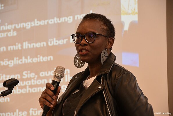 Foto MO VA: eine Frau mit großen Ohrringen und Brille sprechend in ein Mikrofon in der Hand im Hintergrund Text einer Präsentation