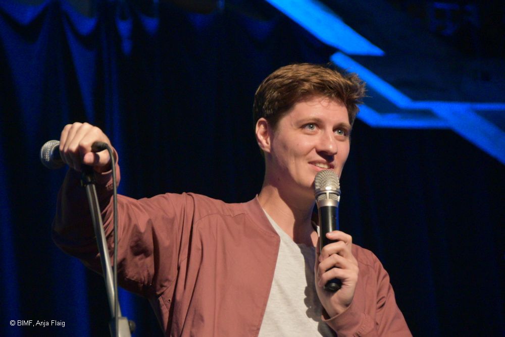 Mann stehend in einer Hand ein Mikrofon, die andere Hand stützt an einem weiteren Mikrofon