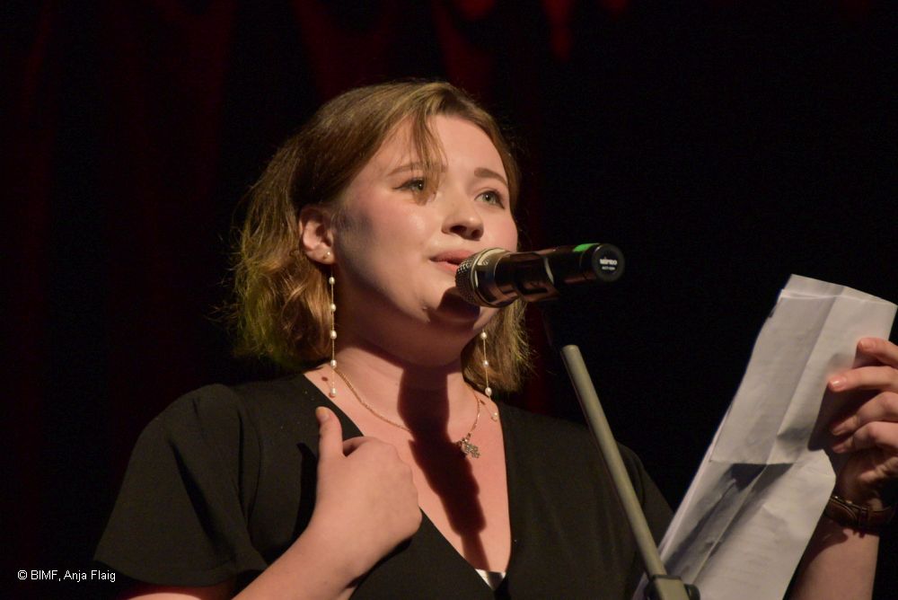 Frau stehend vor einem Mikrofon mit einem Zettel in der Hand, Hintergrund schwarz