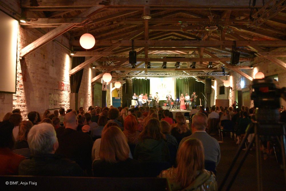 Blick von hinten zur Bühne, viele Menschen sitzend in einem Raum, Beleuchtung