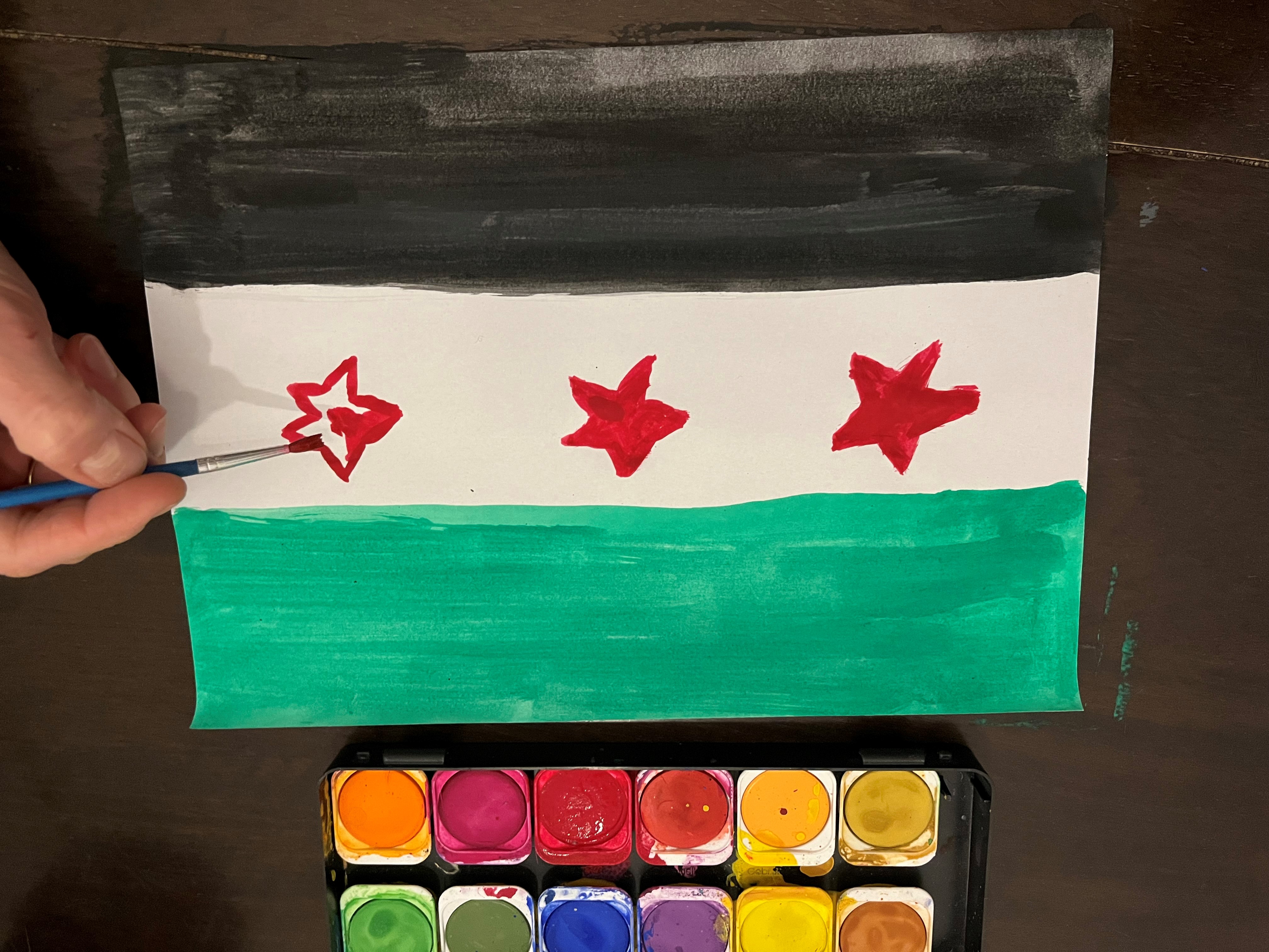 Wasserfarbkasten und Hand, die eine Syrien-Flagge malt: Trikolore in Grün-weiß-schwarz mit drei roten Sternen im mittleren weißen Balken.